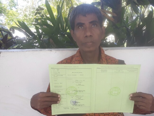 Potret Soleman Matamtasa berdiri di lahan sengketa di Desa Oeteta, Kupang, dengan latar tanaman yang rusak akibat penggusuran.